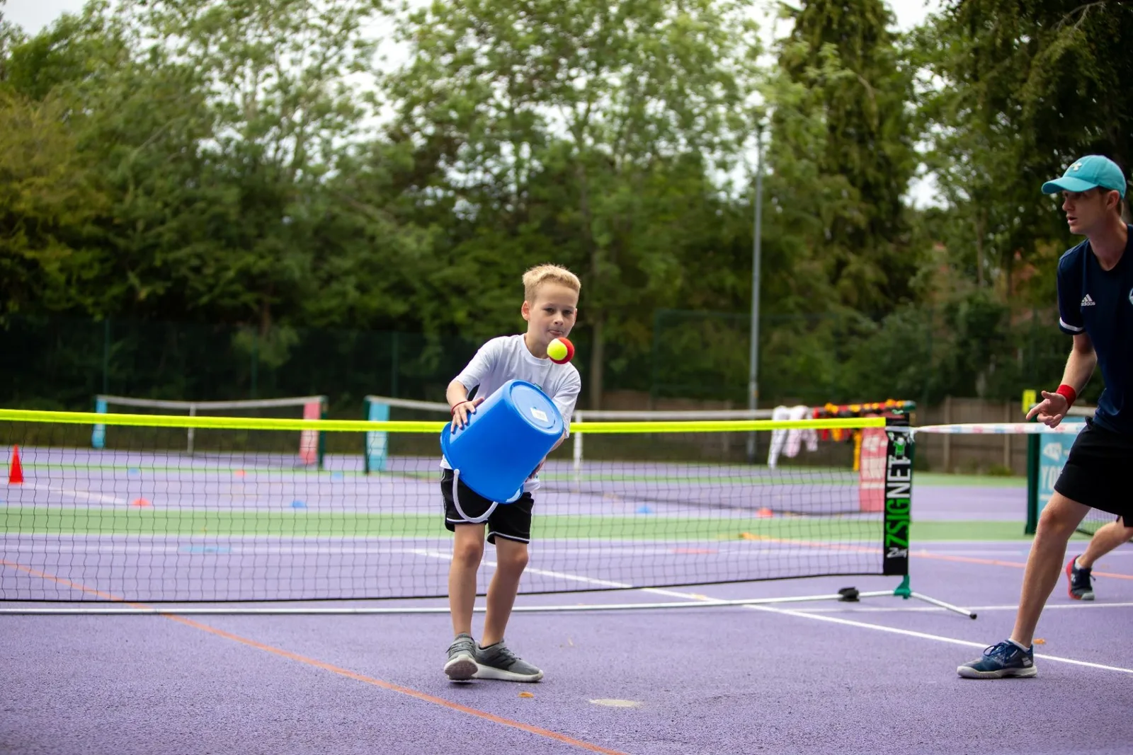 Young player practising a shot with a coach beside the net